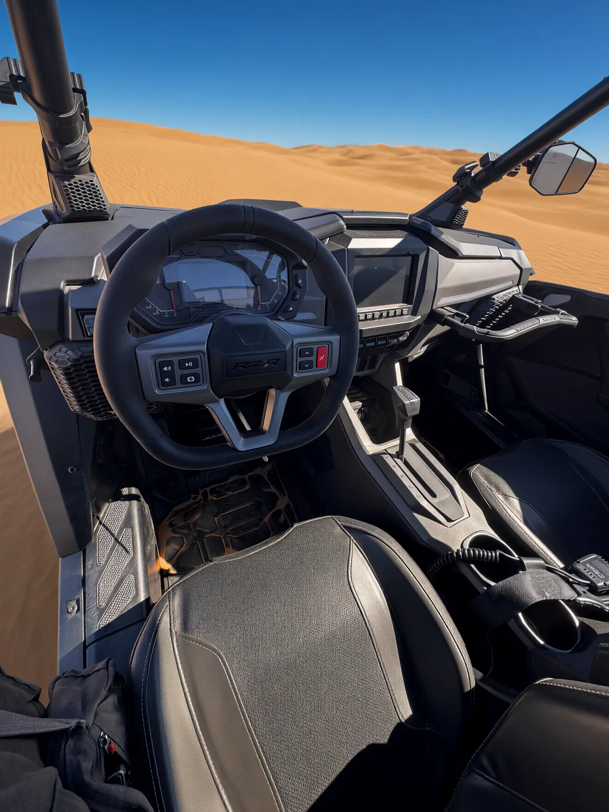 Driver's view of the RZR Pro S Ultimate "Ninja" cockpit with the steering wheel controls, digital gauge, center touchscreen and gear selector, with sand dunes ahead