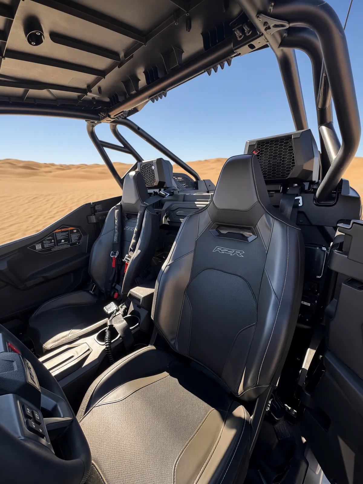 Inside the RZR Pro S Ultimate "Ninja": two RZR bucket seats with harness belts and speaker pods mounted behind the seats under the roll cage, with sand dunes beyond the cabin